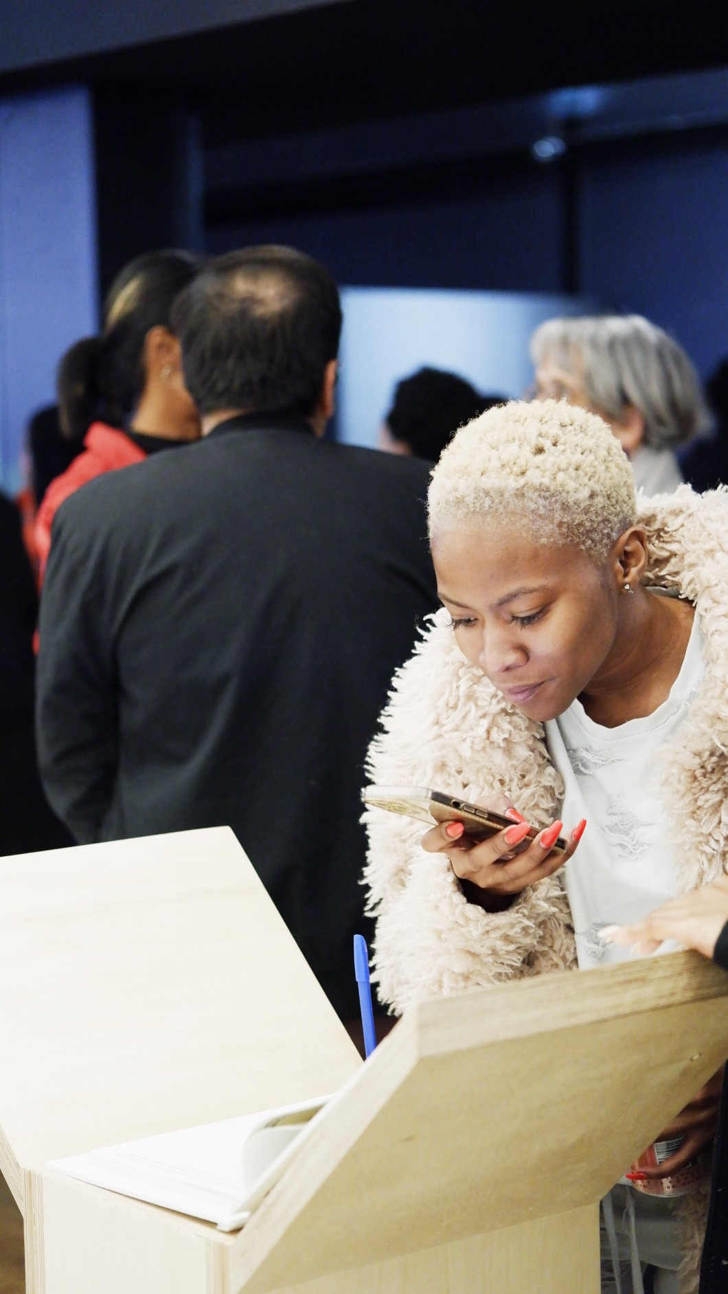 Photo of artwork fragment being engaged with by visitor. Artwork is a series of 6 timber podiums. One podium seen in this photo with woman peeking into podium interior, taking an image with her cellphone.