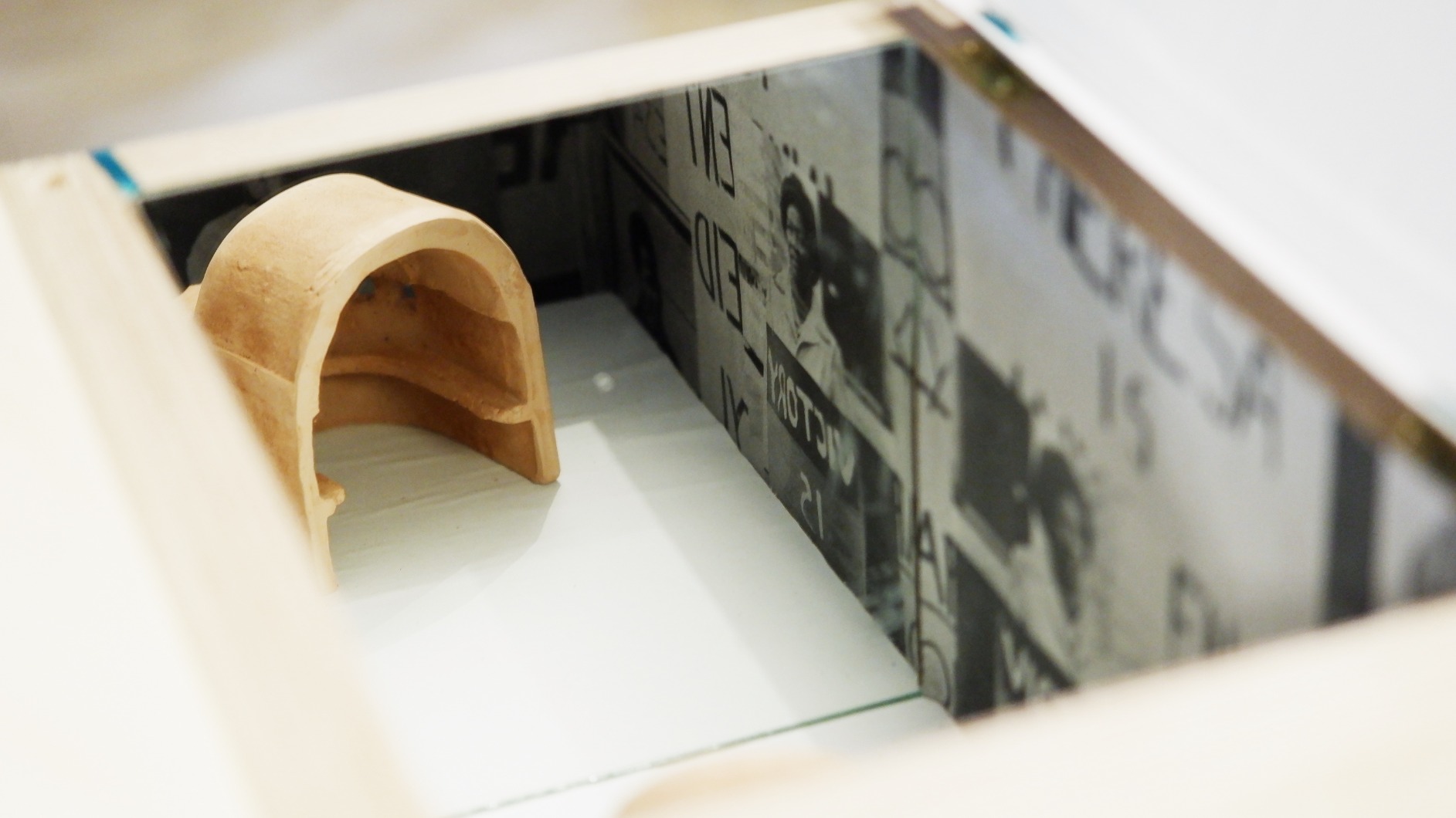 Photo of artwork fragment. Artwork is a series of 6 timber podiums. One podium interior is seen in this photo with collaged faces and a mirror reflecting a clay model of a dome figure.