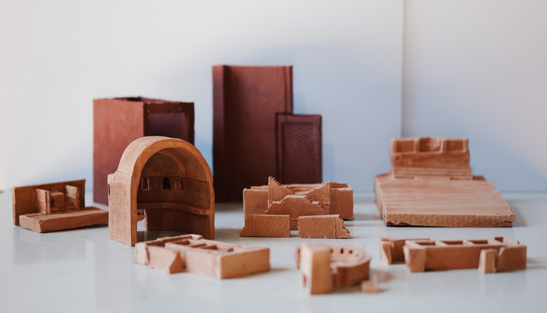 Photo of artwork fragment, showing collection of clay models with white background. The models are abstract figures resembling landscapes, domes, doors. The colours are warm browns.
