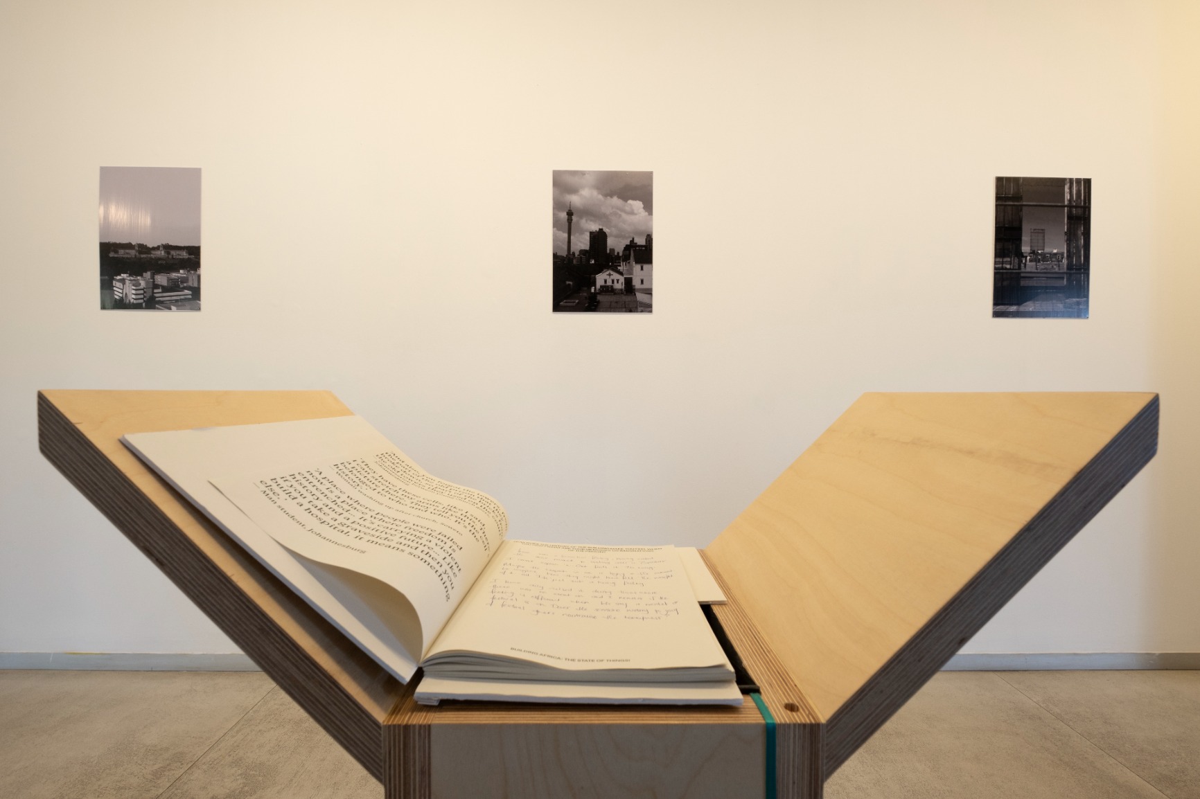 Photo of artwork fragment. Artwork is a series of 6 timber podiums. One podium seen in this photo with a book on the top. Photographs on gallery wall seen in background.