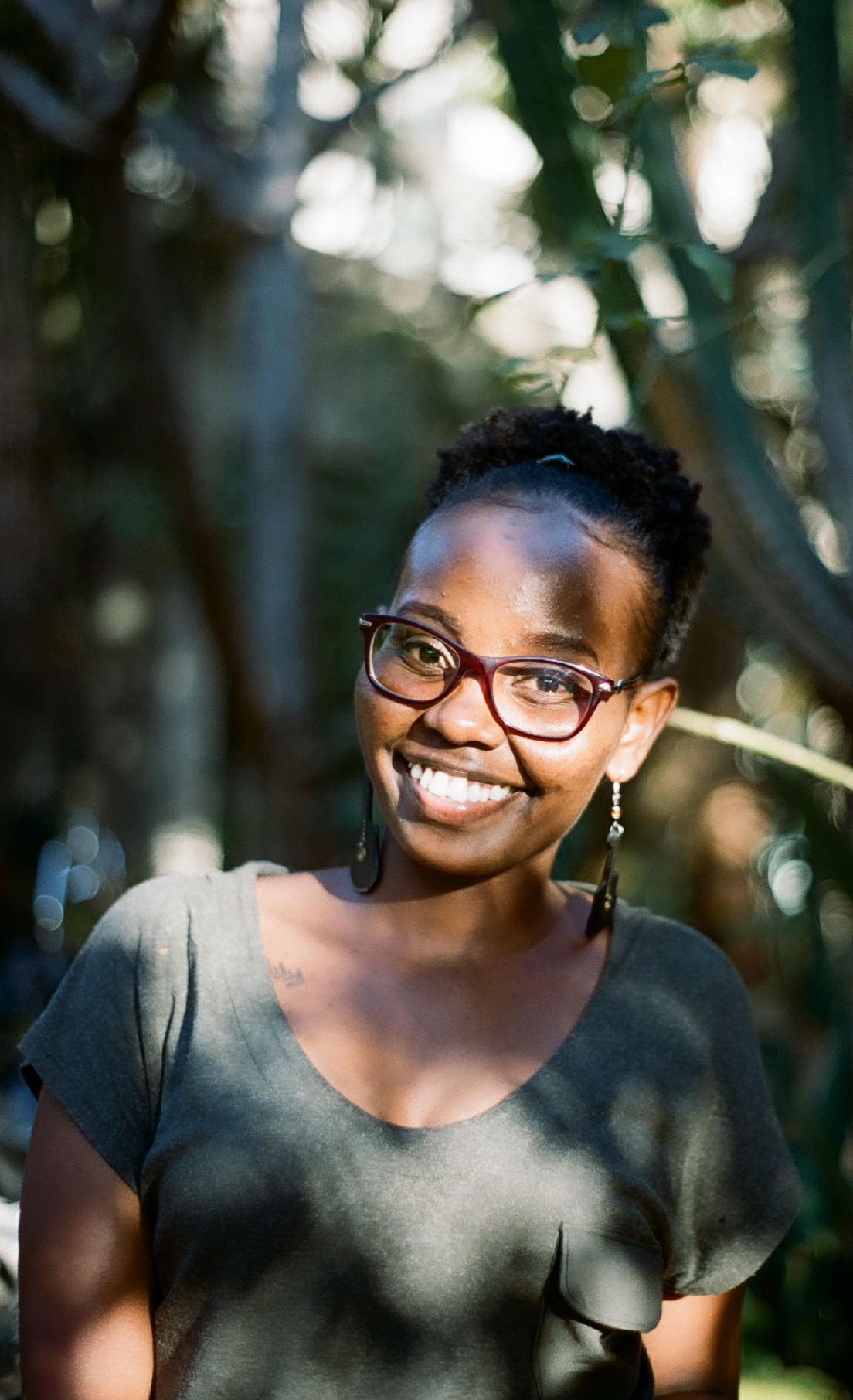 Photo portrait of young African woman smiling with glasses and a green top amongst greenery in the background.