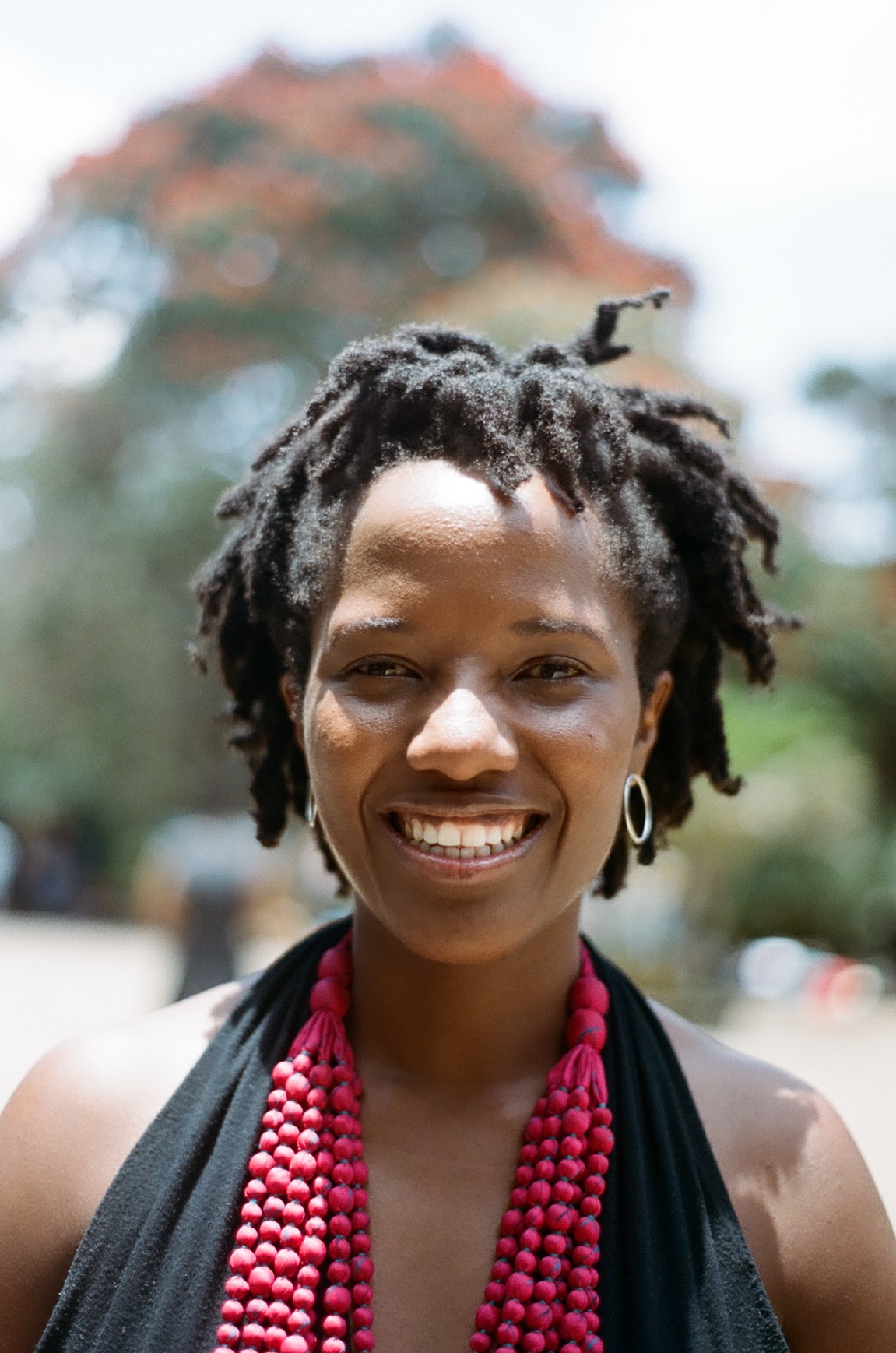 Photo portrait of young African woman smiling with dreads, a black top and a hot pink beaded necklace.