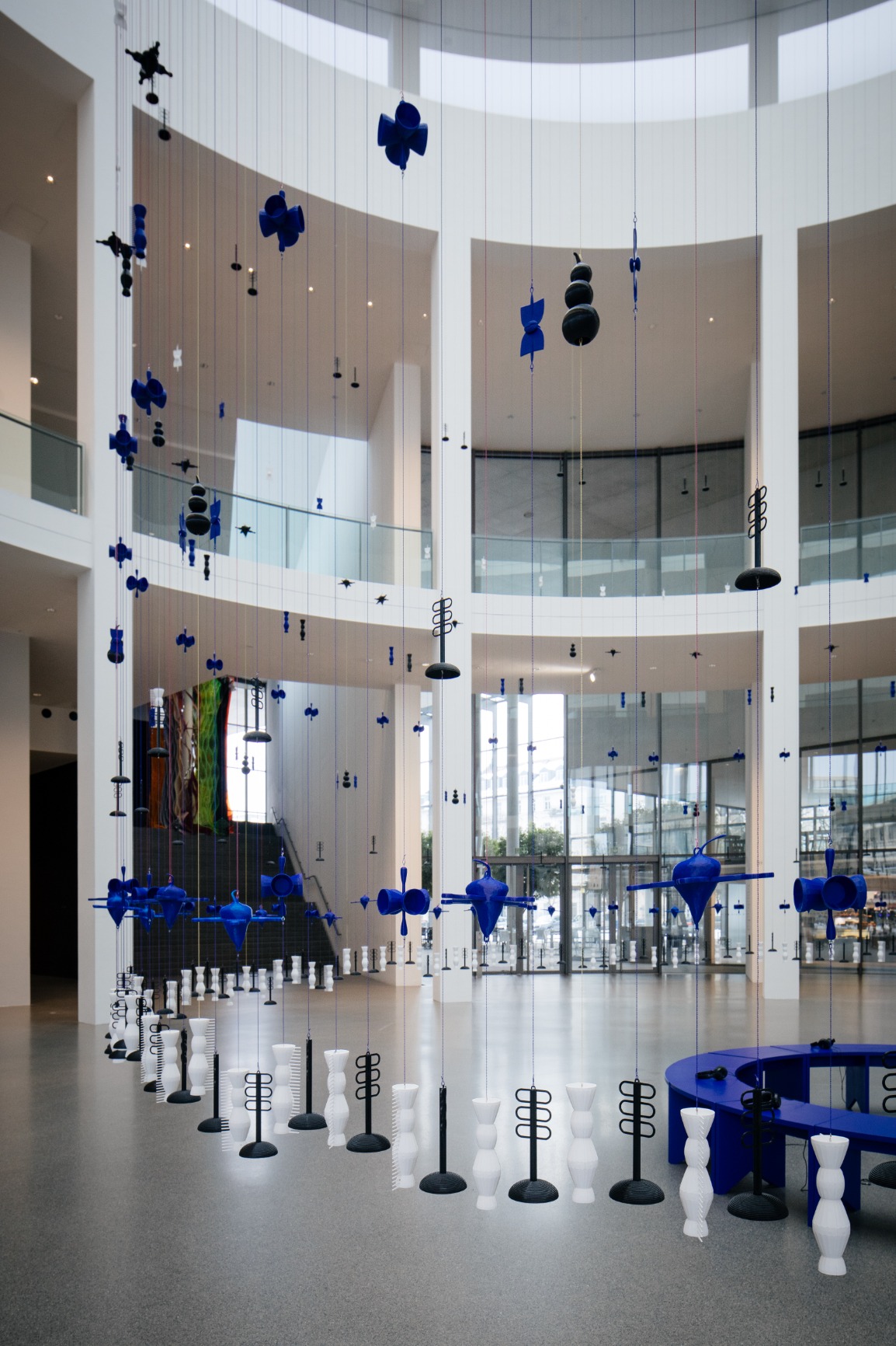 Photo of artwork. Artwork is a transparent beaded hanging curtain of 16m in a circular form creating an interior. The interior has blue benches that when put together form a circle. The curtain beads are blue, white and black. The strings are blue and pink. The image is taken from the side during daylight. The beads are shown in detail.