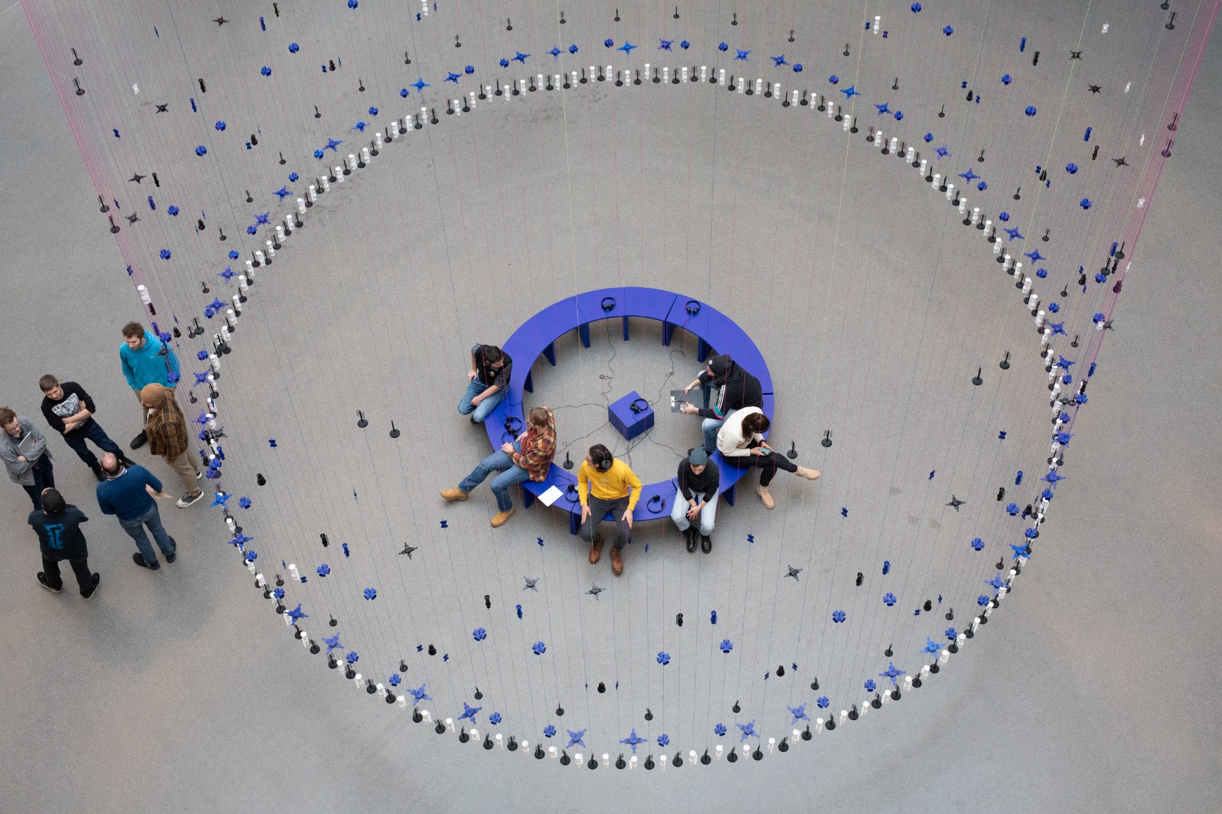Photo of artwork. Artwork is a transparent beaded hanging curtain of 16m in a circular form creating an interior. The interior has blue benches that when put together form a circle. The curtain beads are blue, white and black. The strings are blue and pink. There are people sitting on the bench listening with earphones to a soundscape. There are people standing outside of the curtain. The image is taken from above.