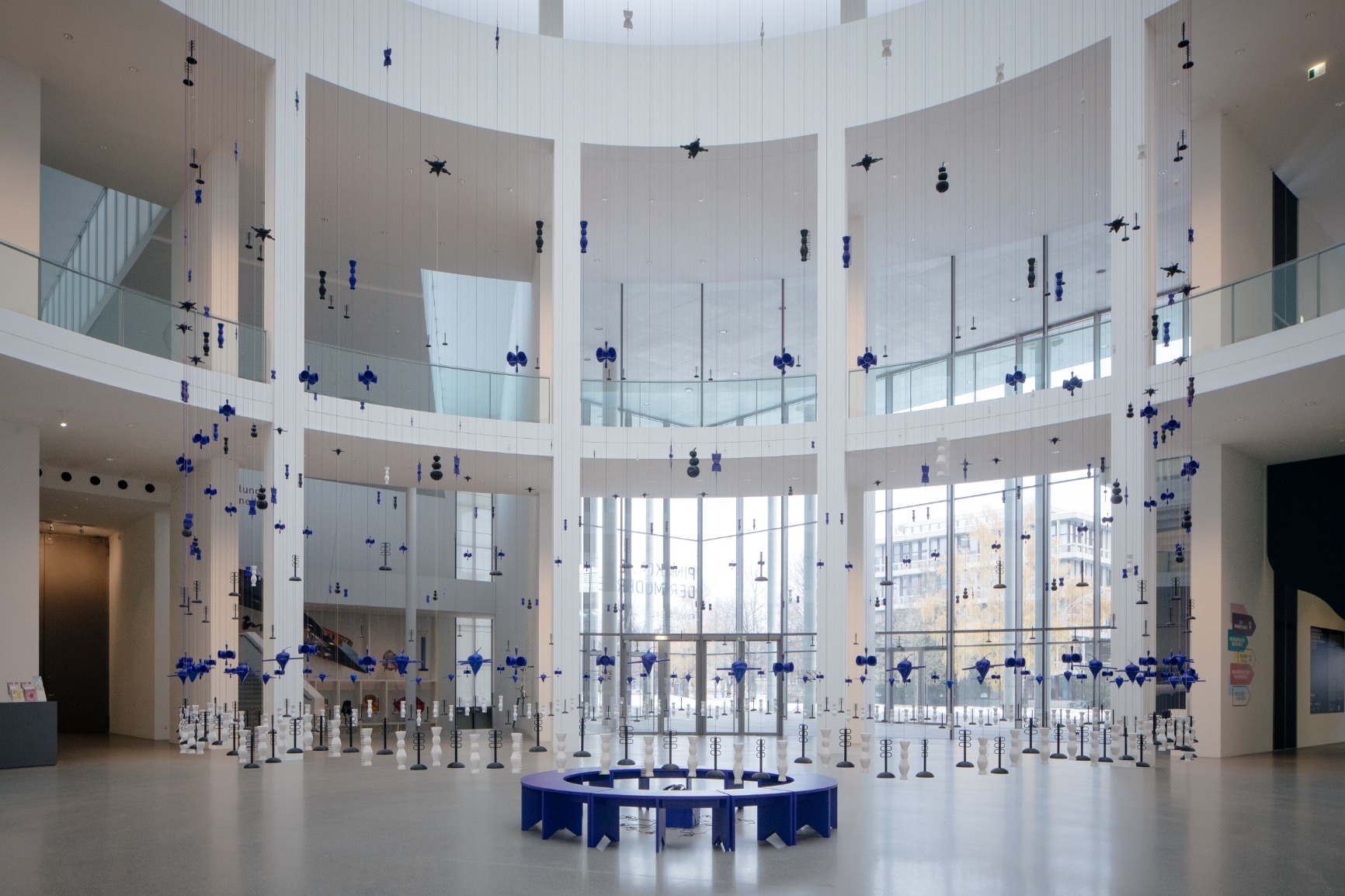 Photo of artwork. Artwork is a transparent beaded hanging curtain of 16m in a circular form creating an interior. The interior has blue benches that when put together form a circle. The curtain beads are blue, white and black. The strings are blue and pink. The image is taken from the side during daylight.