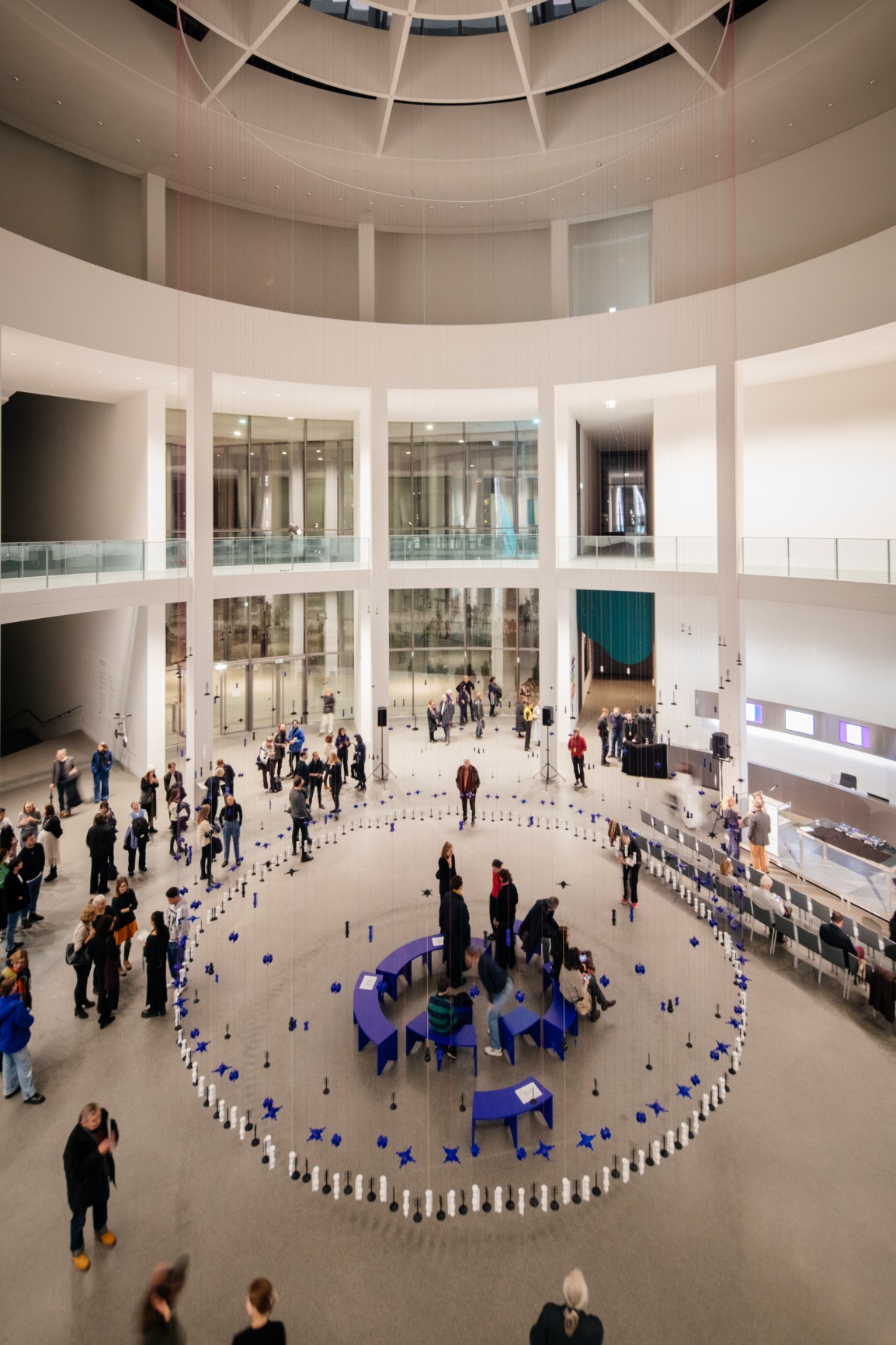 Photo of artwork. Artwork is a transparent beaded hanging curtain of 16m in a circular form creating an interior. The interior has blue benches that when put together form a circle. The curtain beads are blue, white and black. The strings are blue and pink. There are people sitting on the bench listening with earphones to a soundscape. There are people standing outside and inside the curtain. The image is taken during the opening evening.