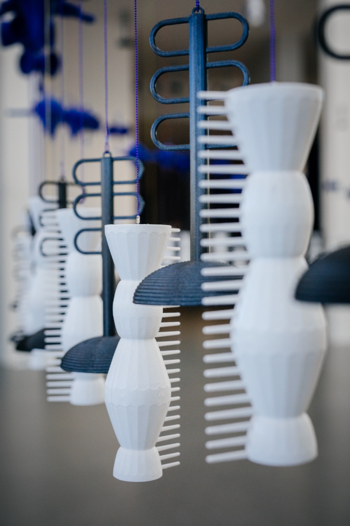 Photo of artwork. The curtain beads are blue, white and black. The strings are blue and pink. The image is taken from the side during daylight. The beads are unique shapes resembles combs, fufu stombers, pap spoons and baskets. The beads are plastic.