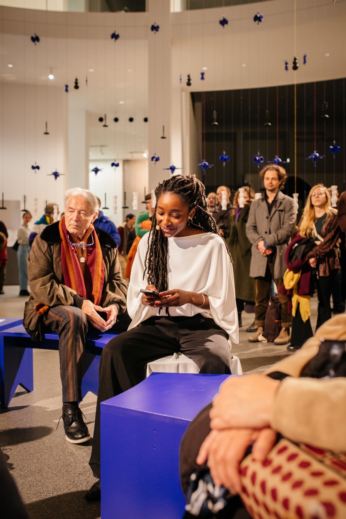 Photo of artwork. Artwork is a transparent beaded hanging curtain of 16m in a circular form creating an interior. The interior has blue benches that when put together form a circle. The curtain beads are blue, white and black. The strings are blue and pink.  There are people standing outside and inside the curtain. The image is taken during the opening evening where Margarida is loudreading.