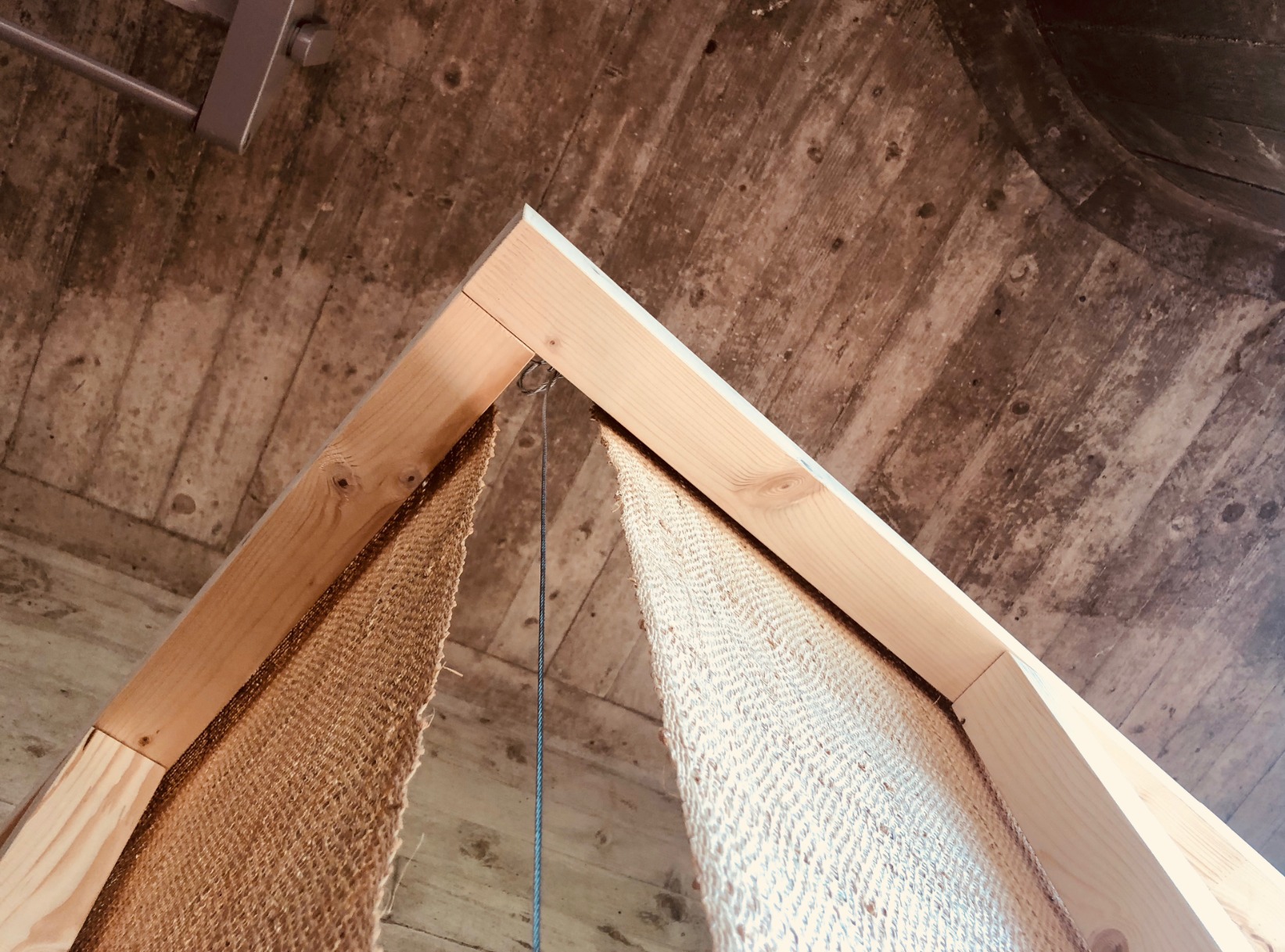 Photo of pavilion detail. Pavilion is simple one-open faced triangular cone. The structural elements are timber. The surfaces are rafia. The pavilion is in an interior space. The colours are warm.