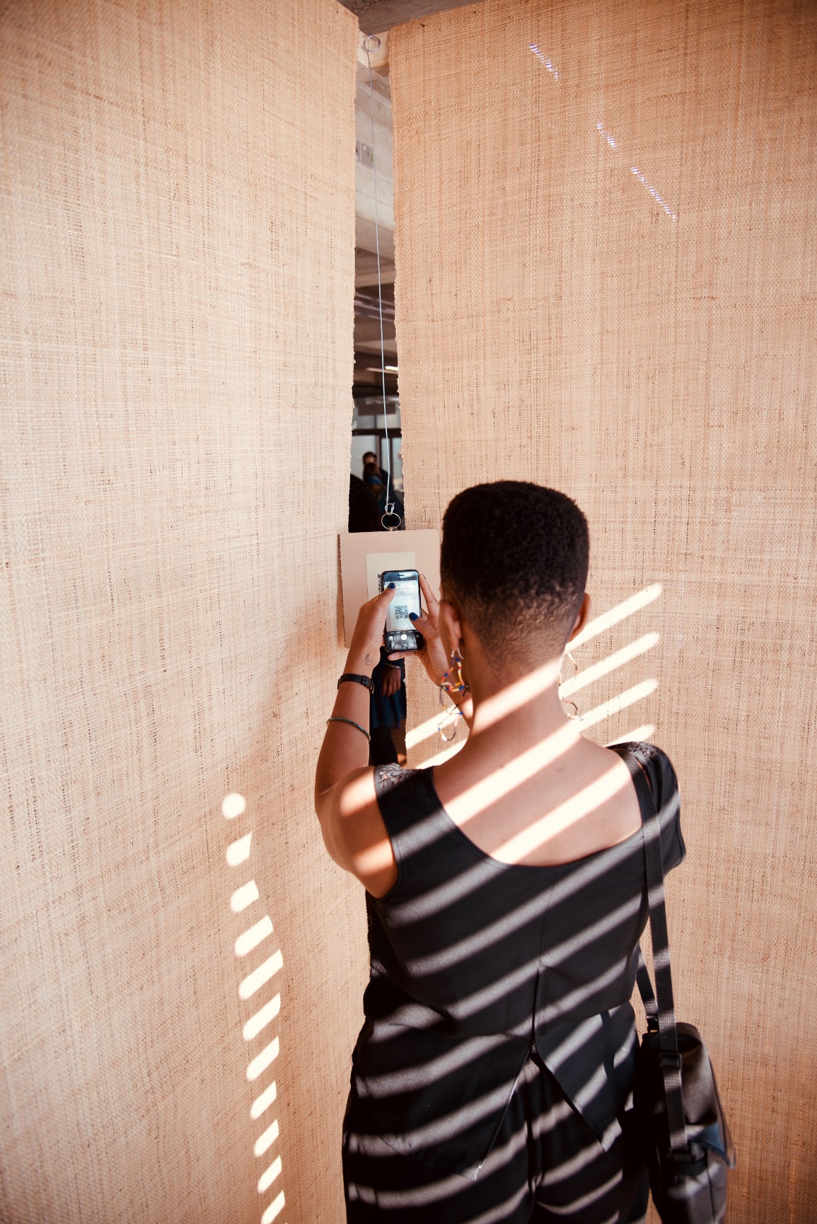 Photo of pavilion with female figure inside scanning a QR code hung in a corner. Pavilion is simple one-open faced triangular cone. The structural elements are timber. The surfaces are rafia. The pavilion is in an interior space. The colours are warm.