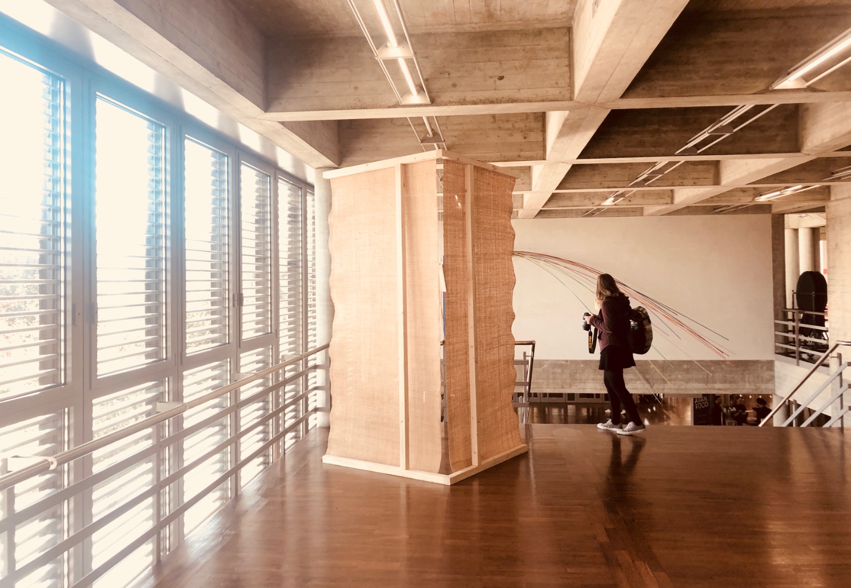 Photo of pavilion. Pavilion is simple one-open faced triangular cone. The structural elements are timber. The surfaces are rafia. The pavilion is in an interior space. The colours are warm.