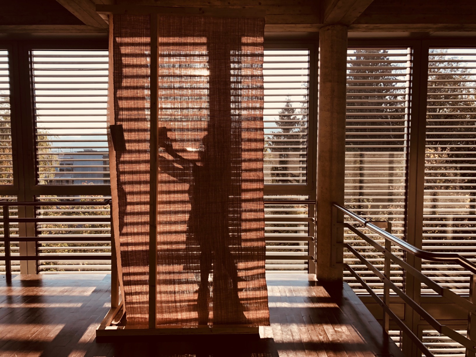 Photo of pavilion and silhouette figure inside space. Pavilion is simple one-open faced triangular cone. The structural elements are timber. The surfaces are rafia. The pavilion is in an interior space. The colours are warm.