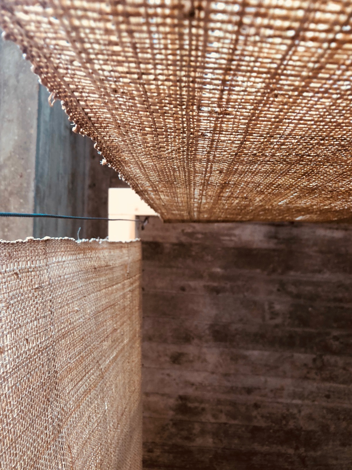 Photo of pavilion. Pavilion is simple one-open faced triangular cone. The structural elements are timber. The surfaces are rafia. The pavilion is in an interior space. The colours are warm.