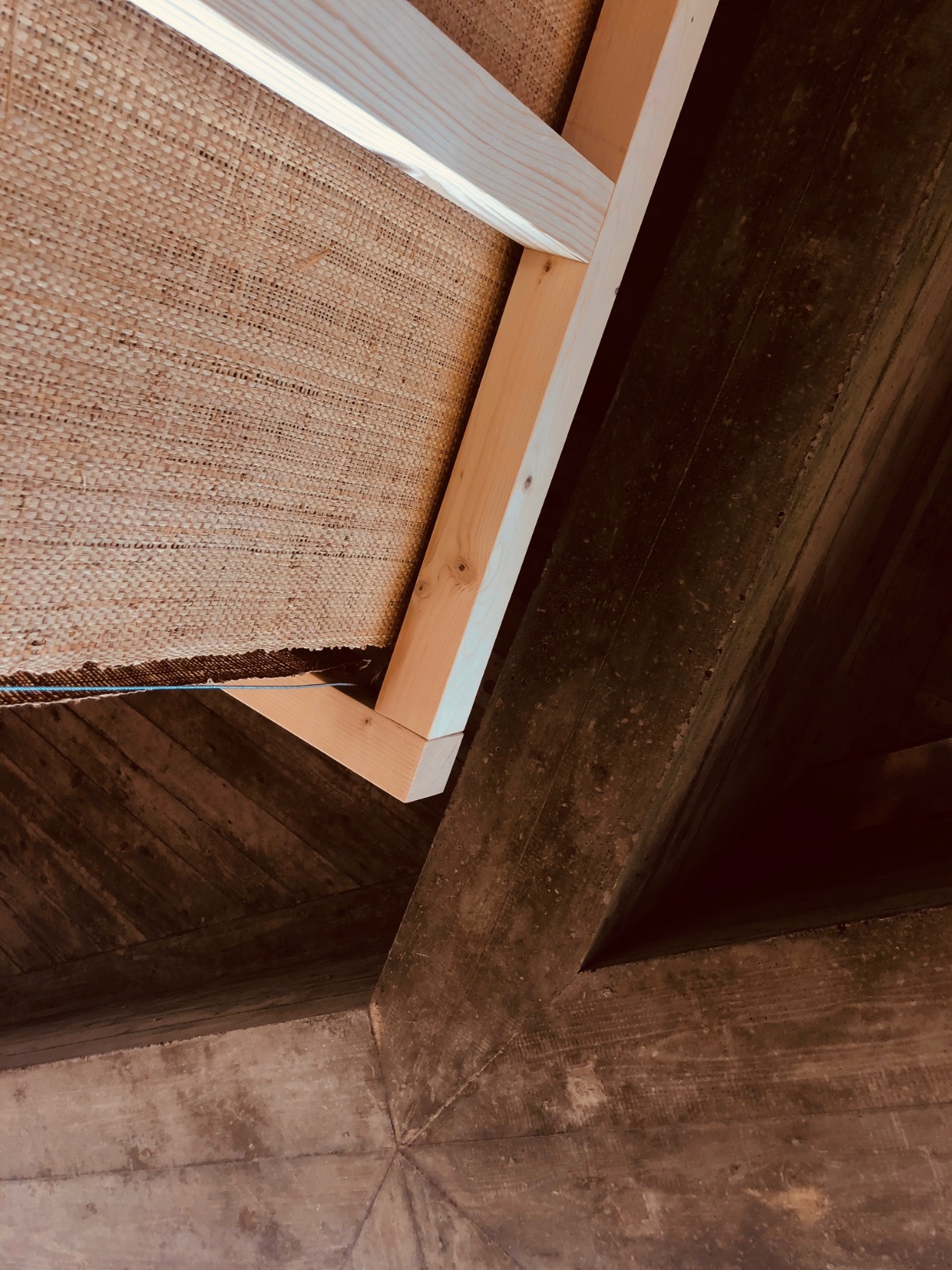 Photo of pavilion. Pavilion is simple one-open faced triangular cone. The structural elements are timber. The surfaces are rafia. The pavilion is in an interior space. The colours are warm.