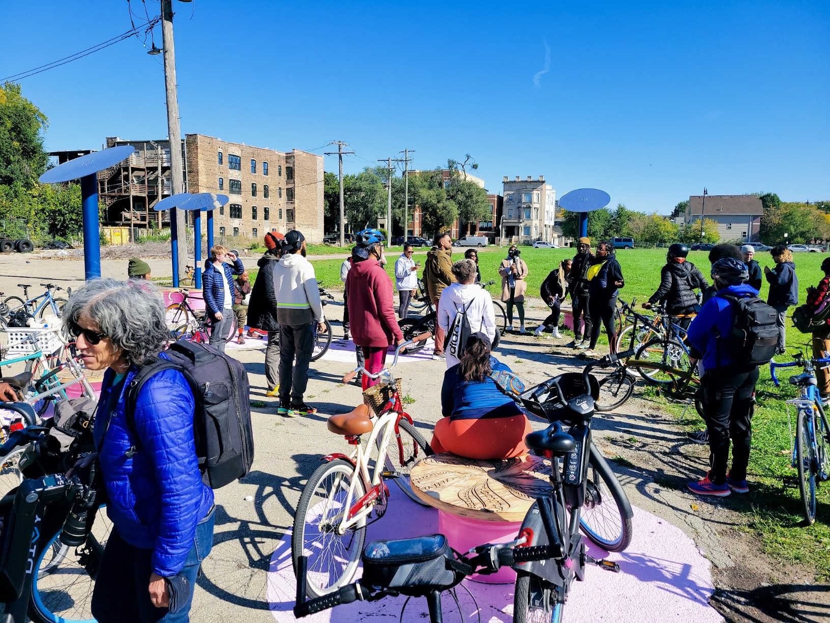 Photo showing pavilion occupied by people. The people are standing amongst their bicyles inbetween the pavilion units. The colours are bright pink, green and the blue sky.