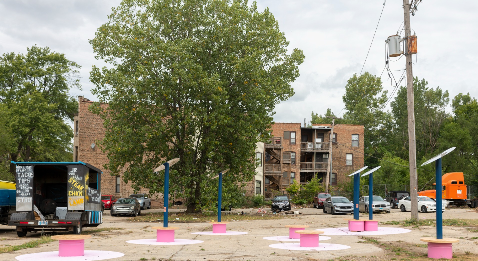 Photo showing pavilion from afar. Reflective disks are held on top of blue poles which are fixed into pink benches with timber tops. The full lawn is visible and a food truck is next to the pavilion.