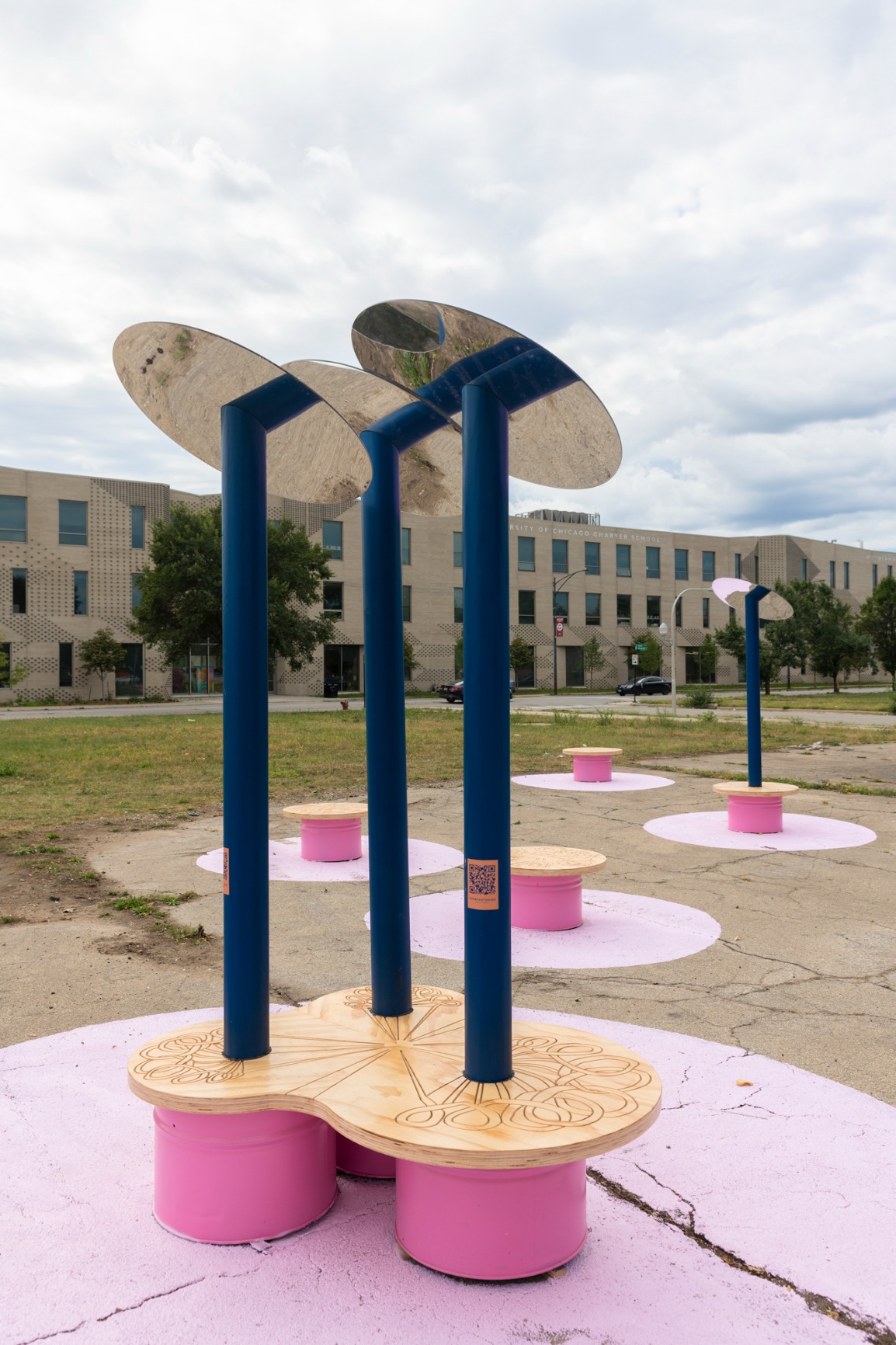 Photo showing pavilion. Reflective disks are held on top of blue poles which are fixed into pink benches with timber tops. There are pink rings painted on the ground where a bench appears. There are QR codes on each pole.