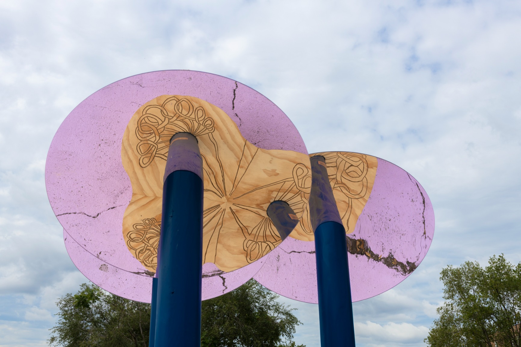 Photo showing pavilion. Reflective disks are held on top of blue poles. The reflection shows a timber organically shaped bench with braid engravings on it. The sky is visible.