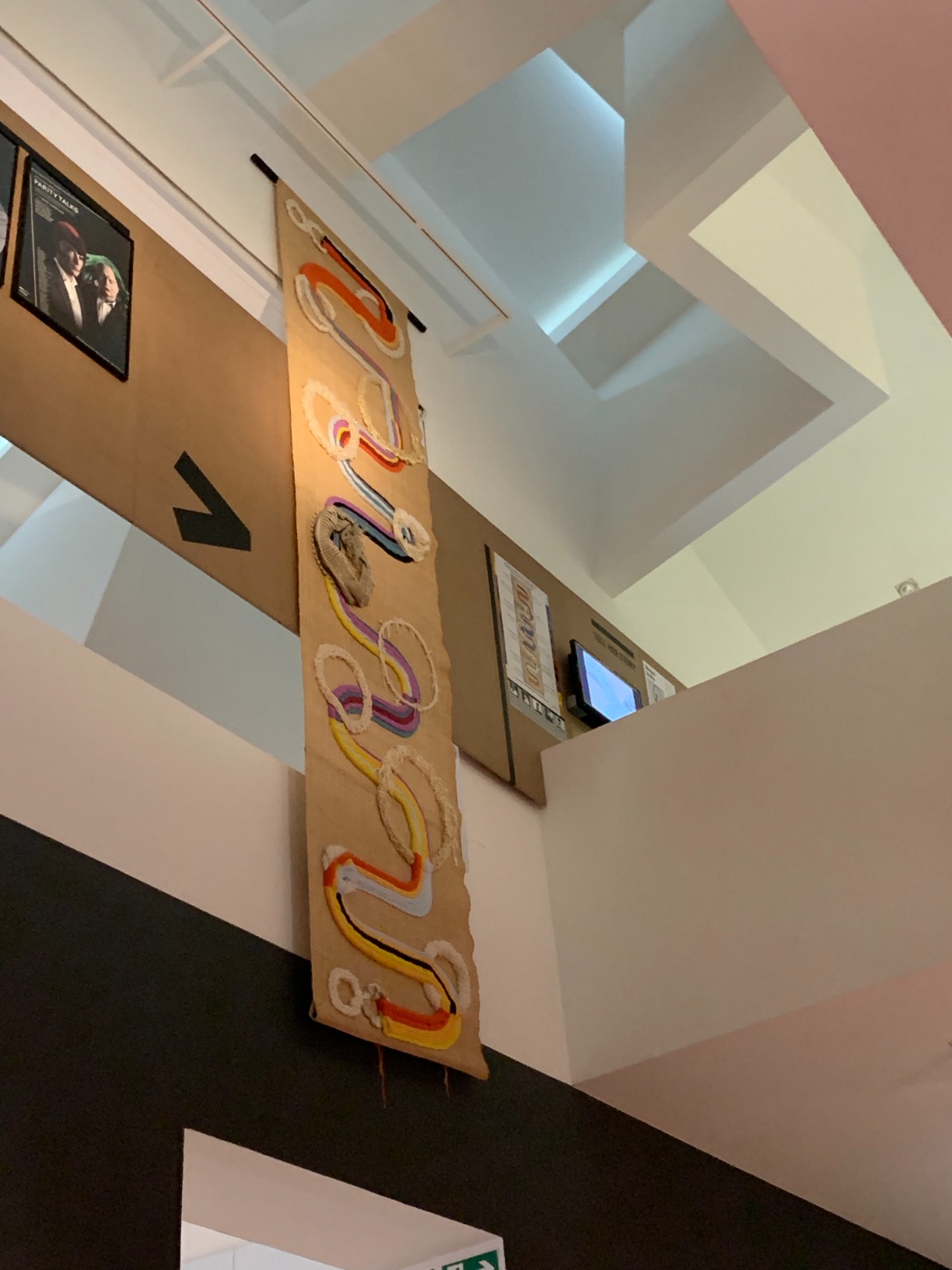 Photo of artwork (colourful tapestry) hung from the ceiling, next to a poster explaining the colour-coded artwork as well as a soundscape video seen on the screen on the wall.