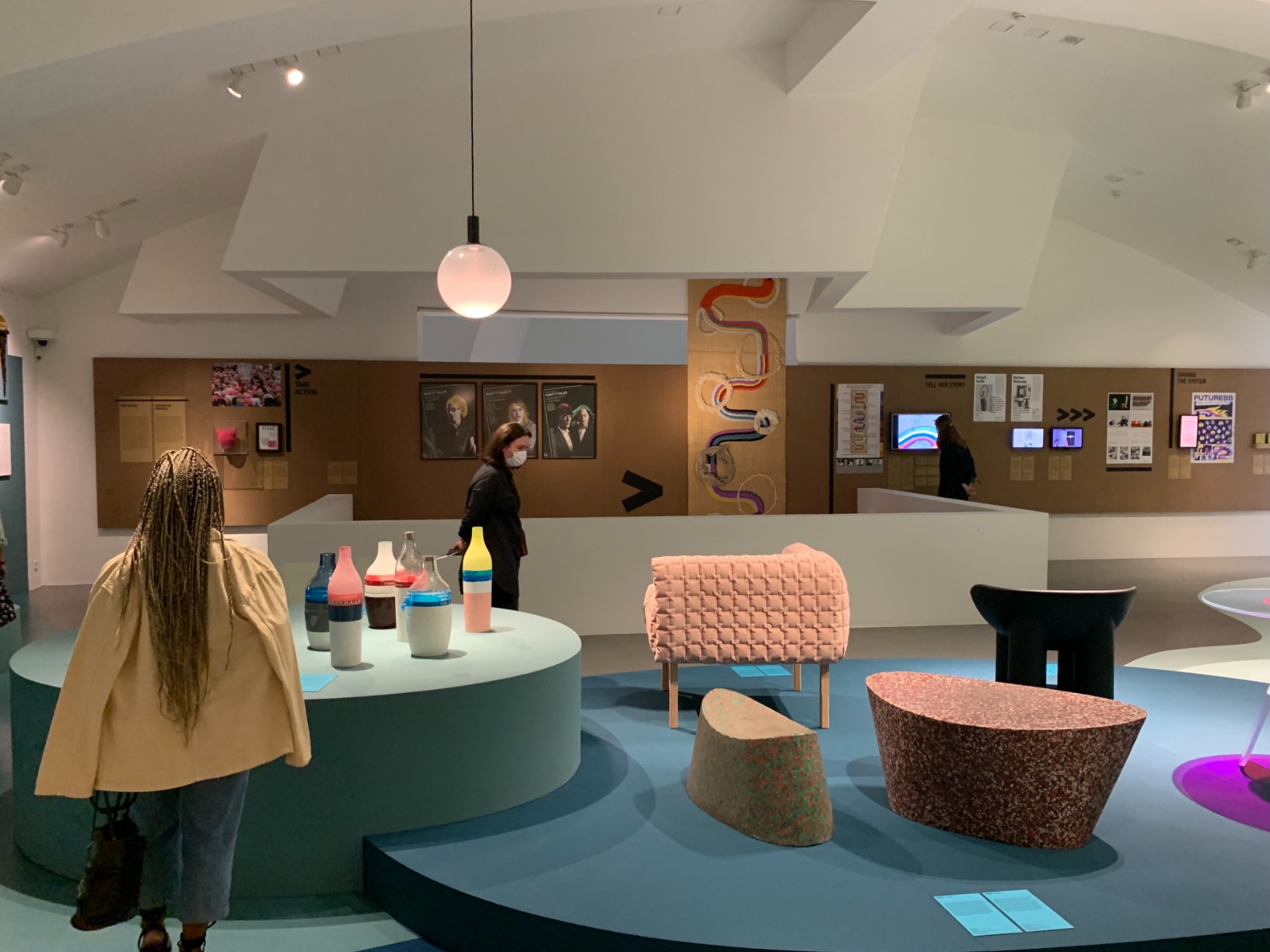 Photo of artwork (colourful tapestry) hung from the ceiling amongst the other artworks in the group show present in the room including furniture objects, and paper-based items on the walls