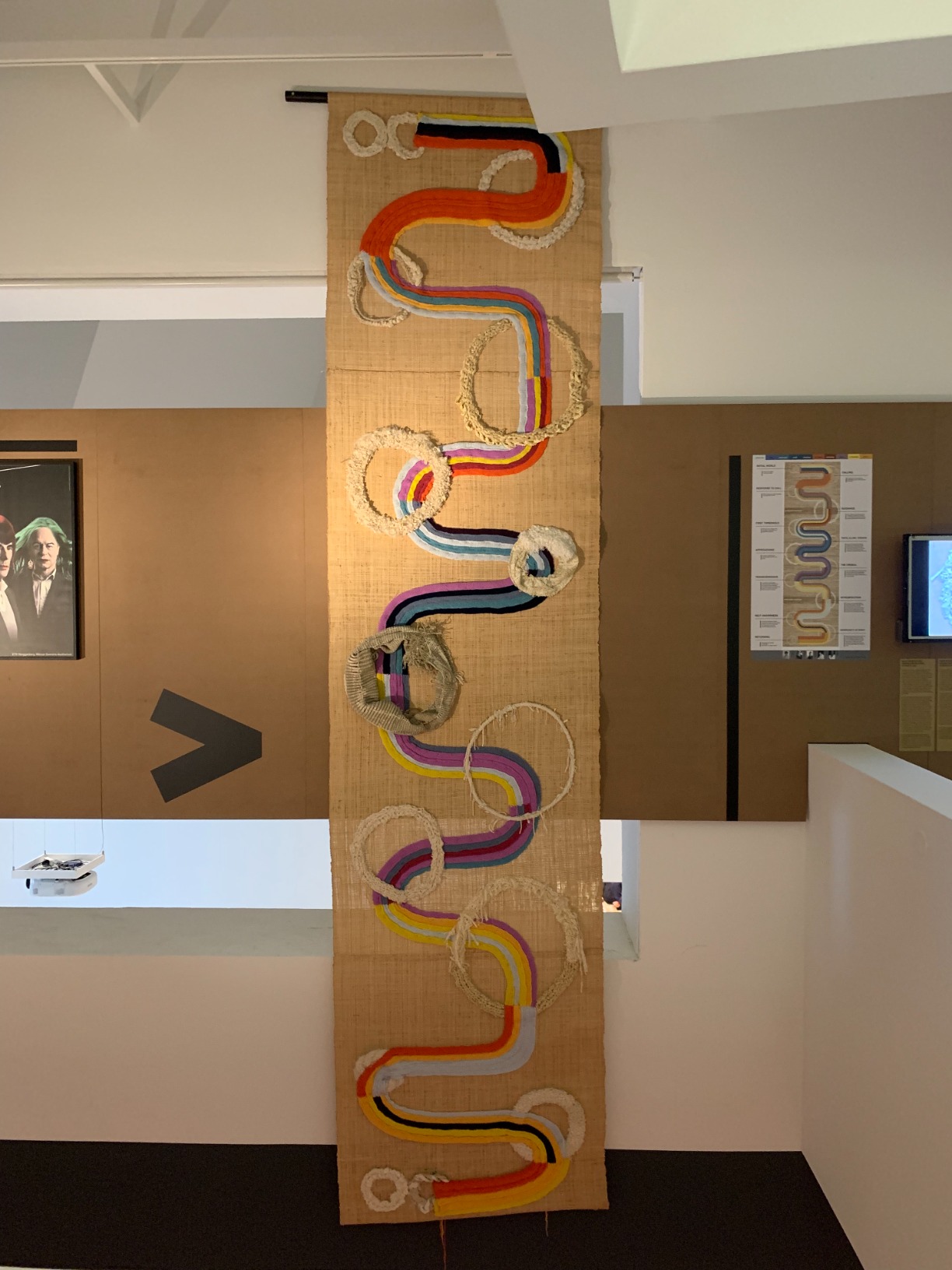 Photo of artwork (colourful tapestry) hung from the ceiling, next to a poster explaining the colour-coded artwork as well as a soundscape video seen on the screen on the wall.