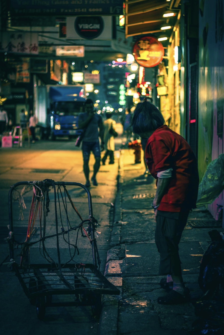 Image of old woman standing in vulnerable stance on the sidewalk in Hong Kong looking down the street next to a wheelable trolly